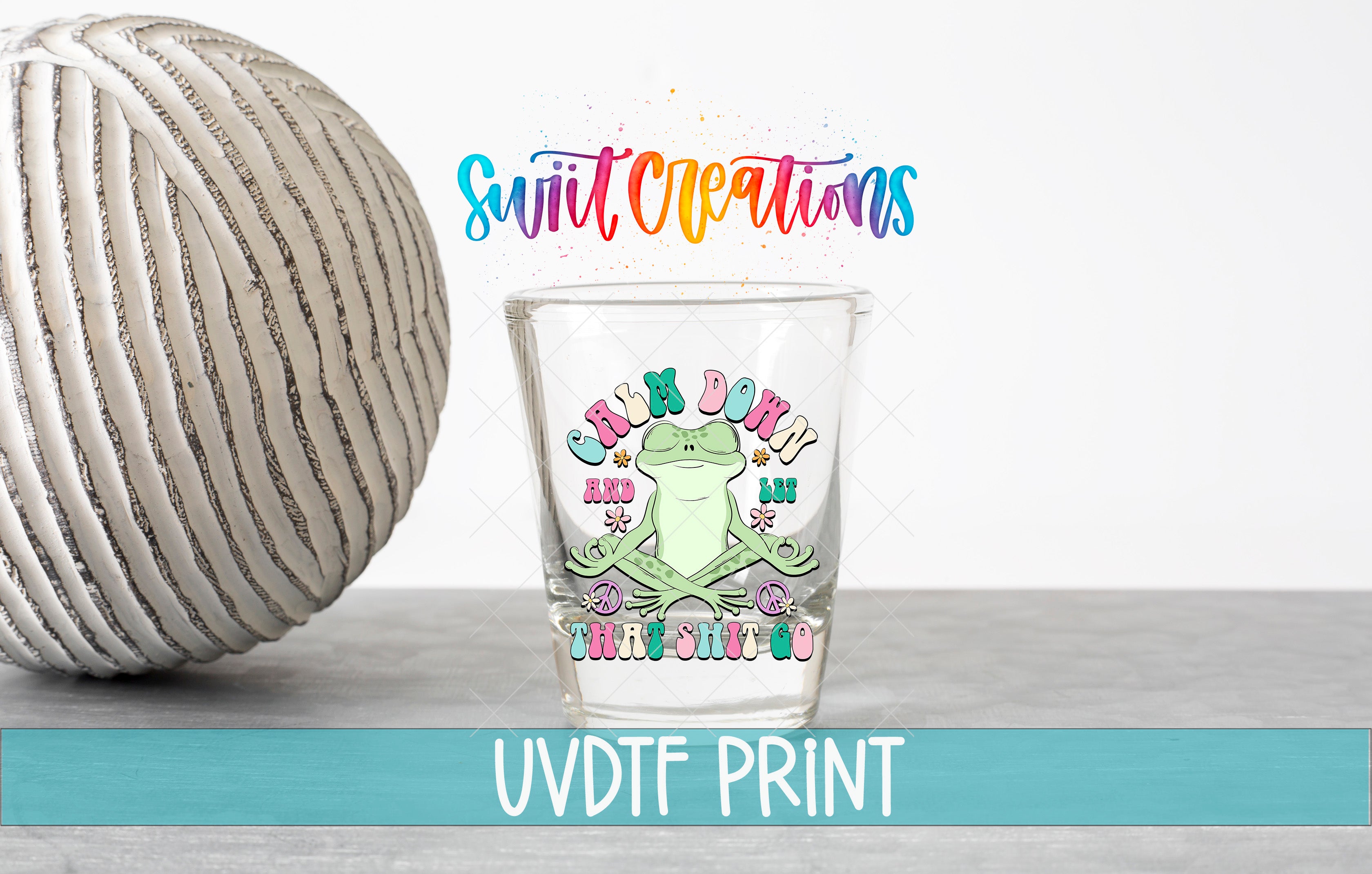 a glass with a frog design and text, placed next to a white ball on a gray surface.