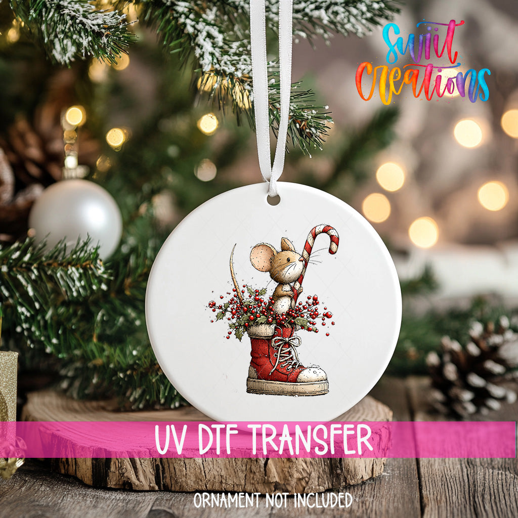A white ornament with a mouse and a candy cane is hanging on a Christmas tree, with a pink ribbon around it.