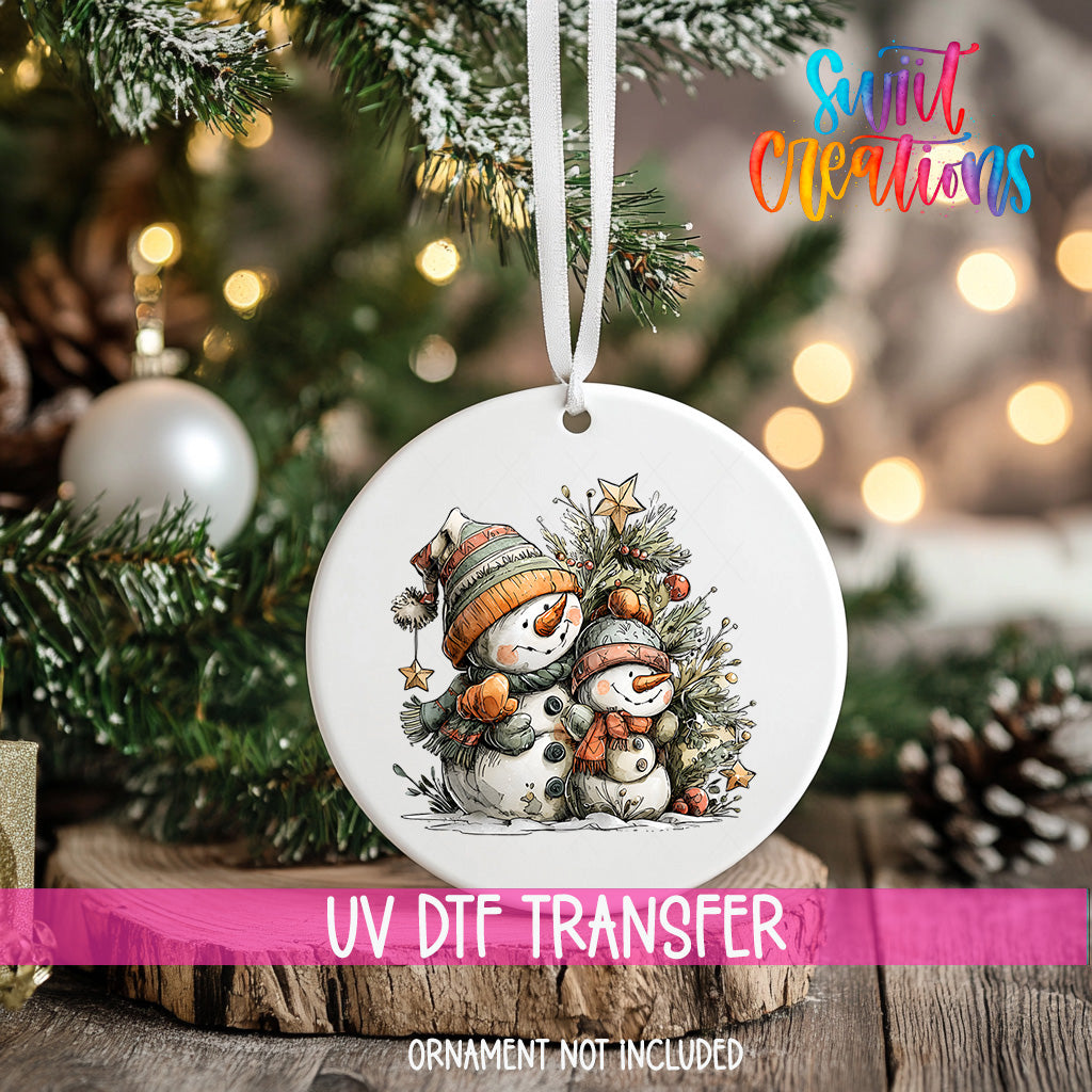 A white ornament with a snowman and star on a Christmas tree, with a pink ribbon around it.