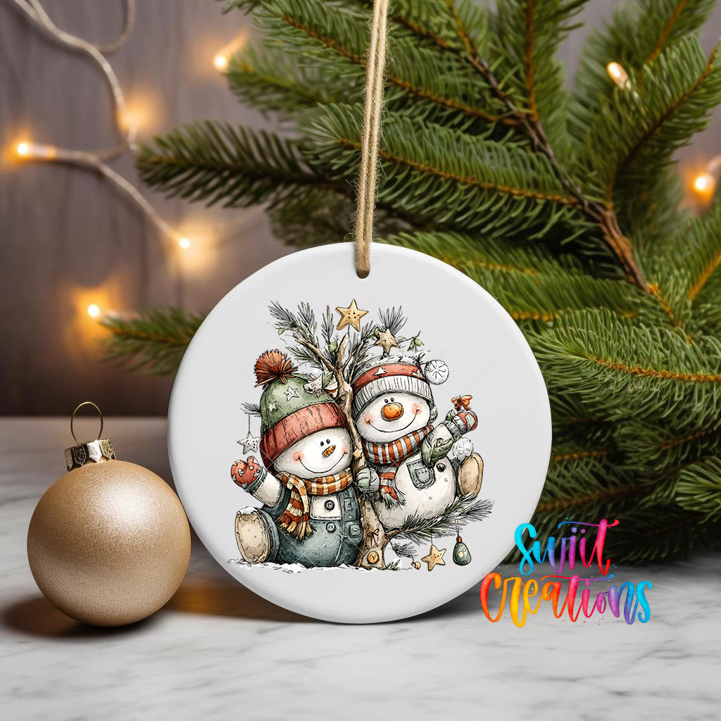 A white ceramic ornament featuring two snowmen, one wearing a green hat and the other a red hat, sitting on a branch with pine needles and a star. The ornament is hanging from a string, and there is a gold ornament next to it on a table. The background includes a Christmas tree with twinkling lights.