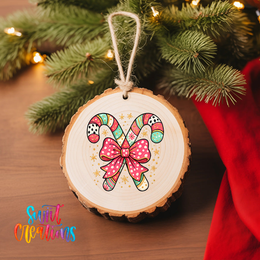 A wooden ornament with a candy cane design and a pink bow is hanging on a Christmas tree, surrounded by greenery and twinkling lights.
