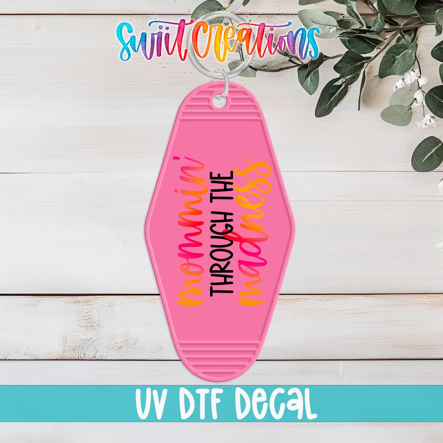 A pink keychain with the text " mommy through the messiness" printed on it, hanging on a wooden surface with some greenery in the background.