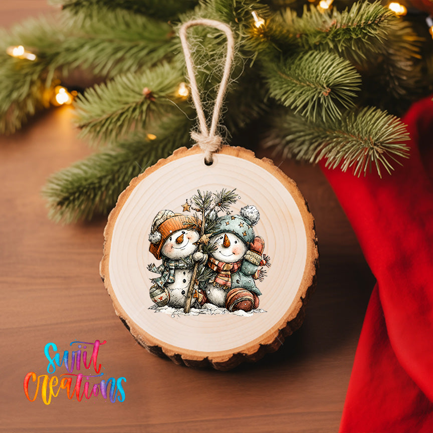 A wooden ornament featuring two snowmen is hanging on a Christmas tree, surrounded by pine branches and twinkling lights.