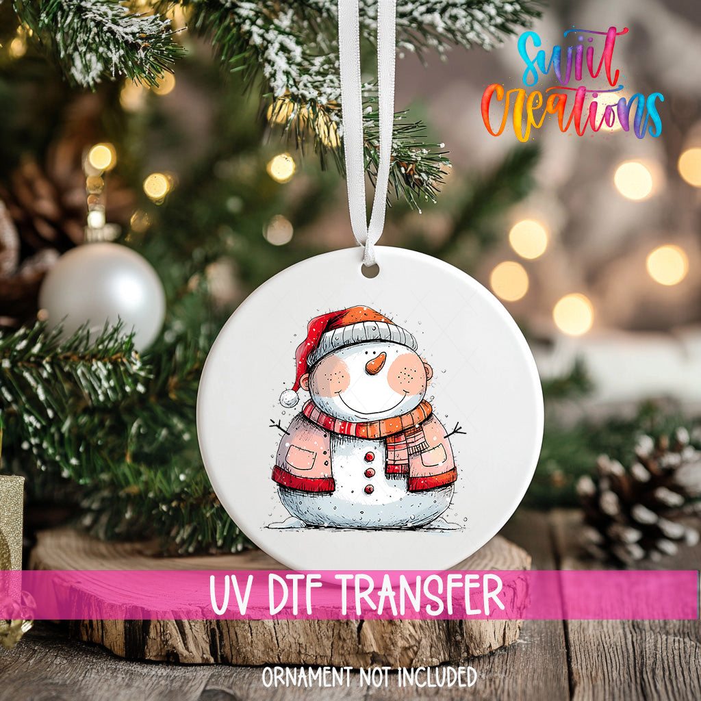 A white ornament with a snowman design hangs on a Christmas tree, surrounded by pine branches and twinkling lights.