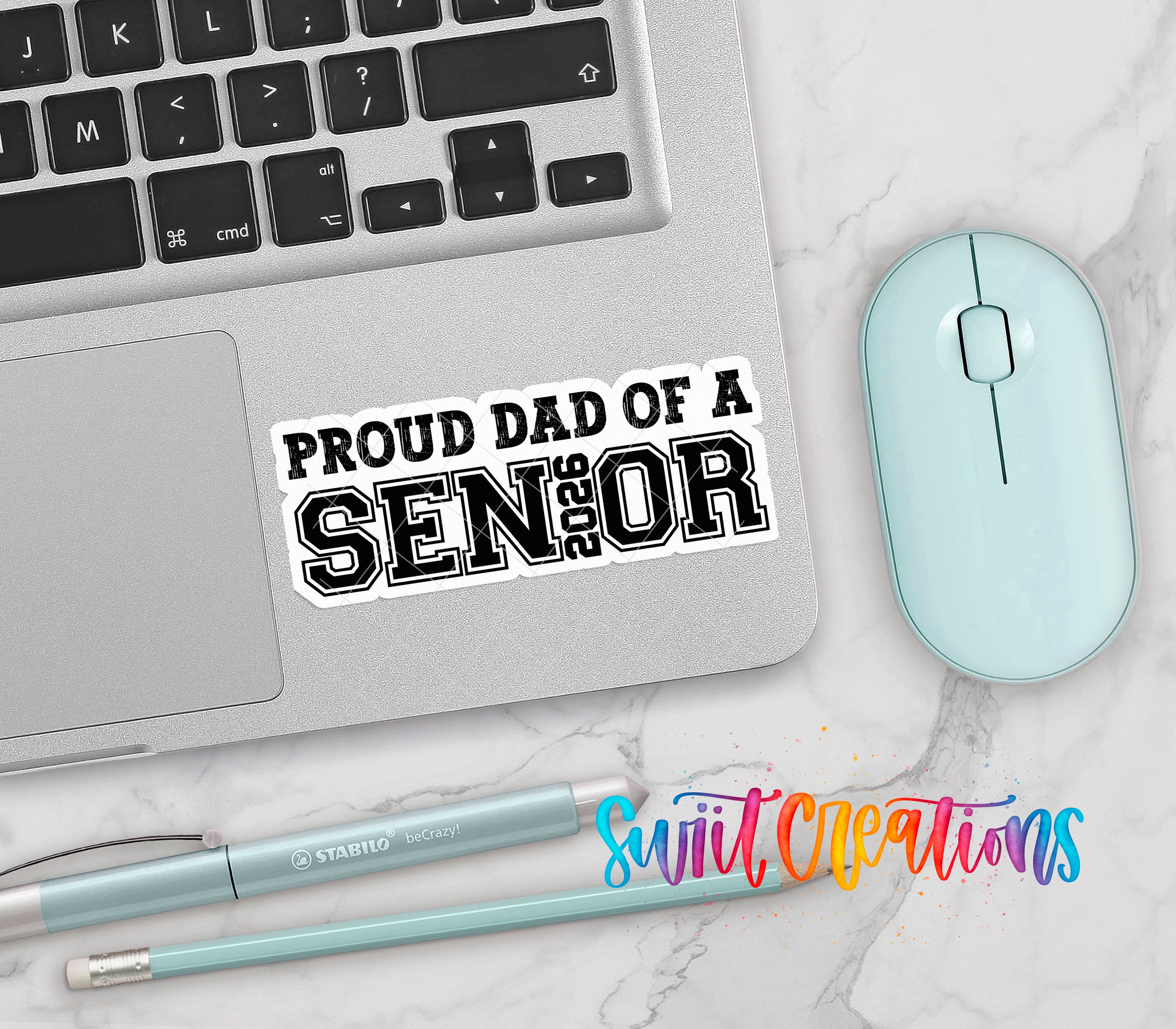 a laptop, a mouse, and various office supplies such as pens and pencils, all placed on a marble surface. There is a sticker on the laptop that says "Proud Dad of a SENIOR".