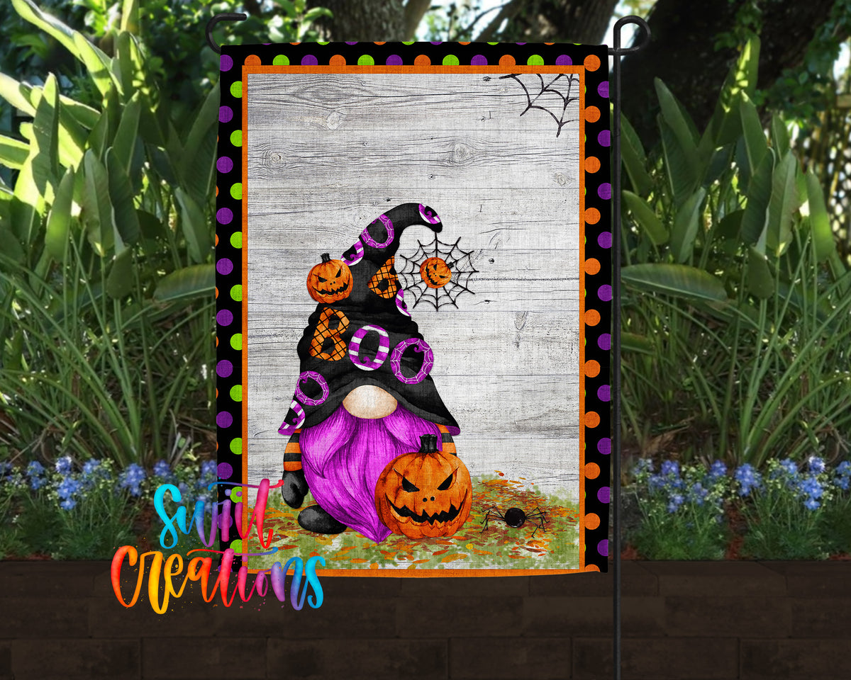 A Halloween-themed garden decoration featuring a gnome with a pumpkin hat and a spider web, surrounded by greenery and flowers.