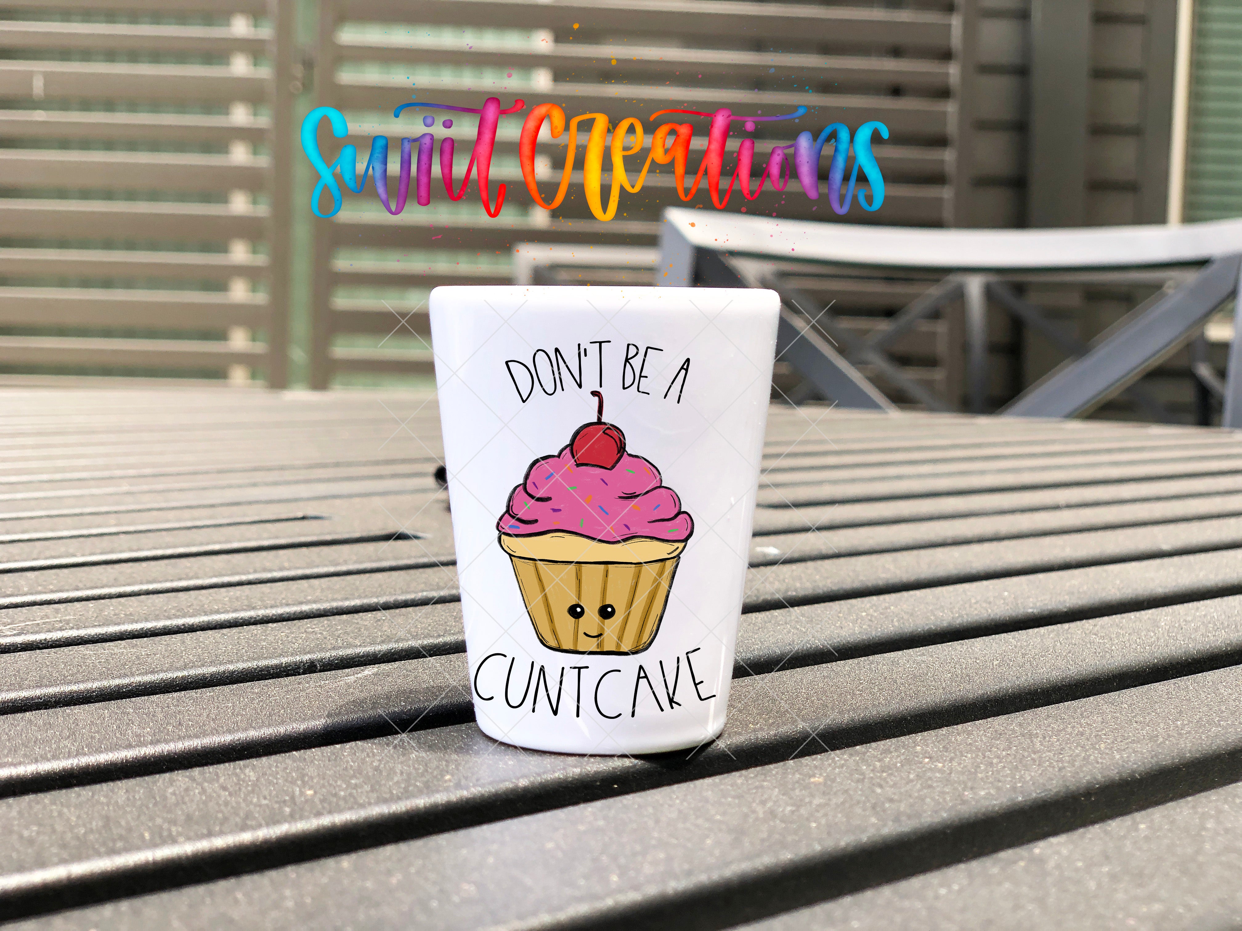 A white cup with a cupcake design and the text "DON'T BE A CUNTCAVE" printed on it, placed on a wooden surface.