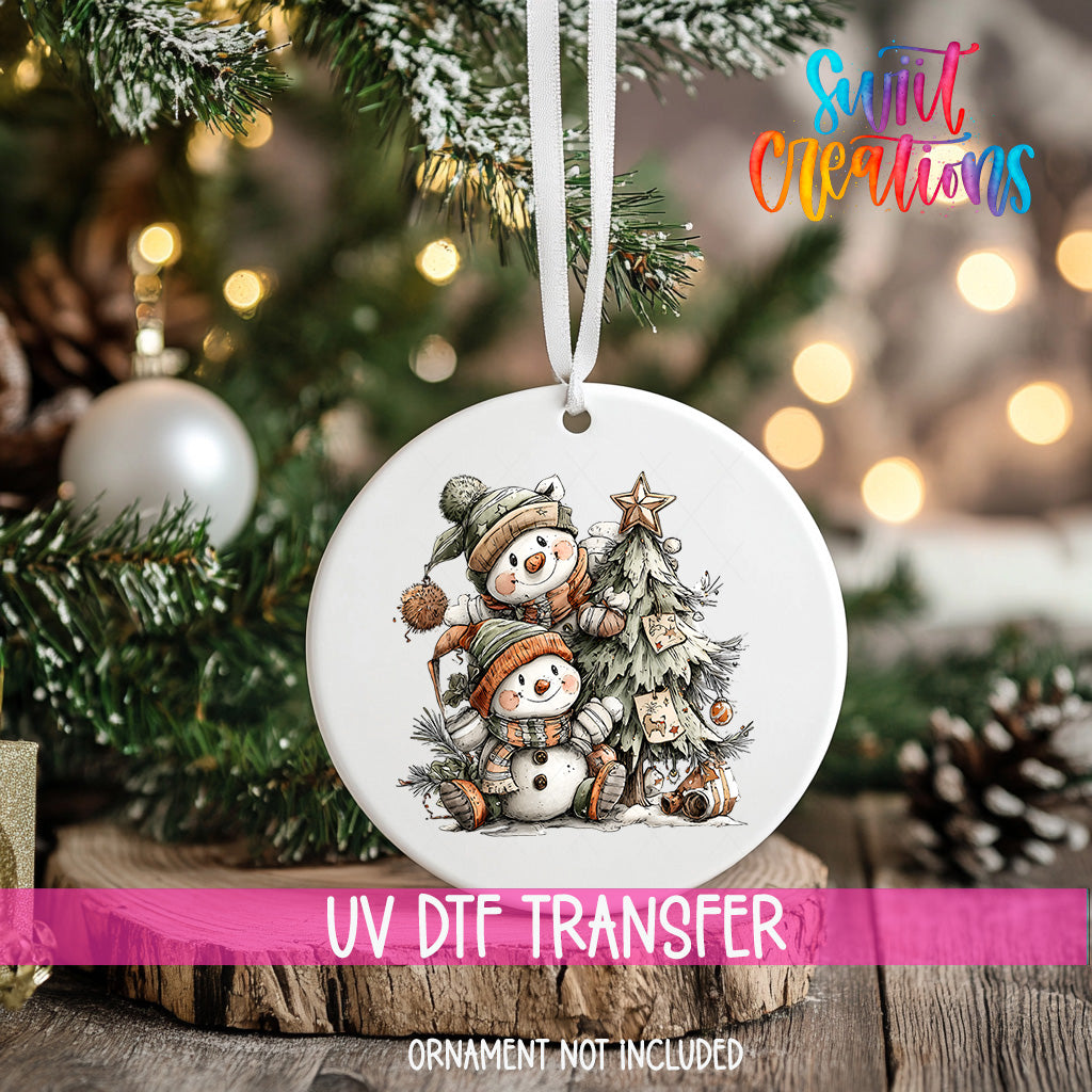 A white Christmas ornament with a snowman and star on a tree, hanging on a tree branch with a blurred background of lights.