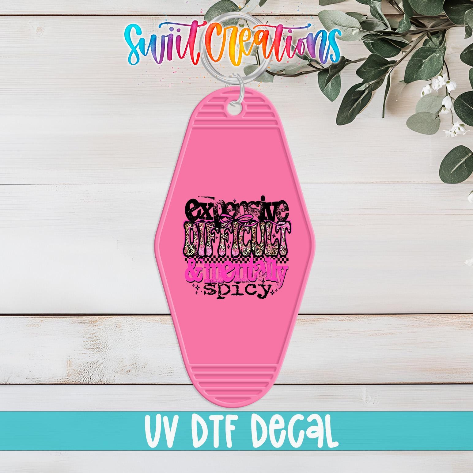 A pink keychain with a decorative design and text on it, placed on a wooden surface with some greenery in the background.