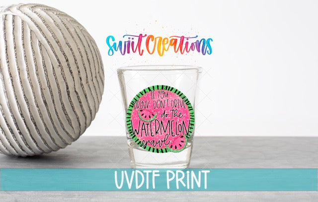 a shot glass with a watermelon-themed design and text, placed next to a white ball on a gray surface.