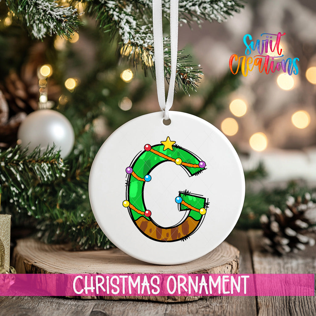 A white Christmas ornament with a green and orange design hangs from a tree branch, with a blurred background of lights and greenery.