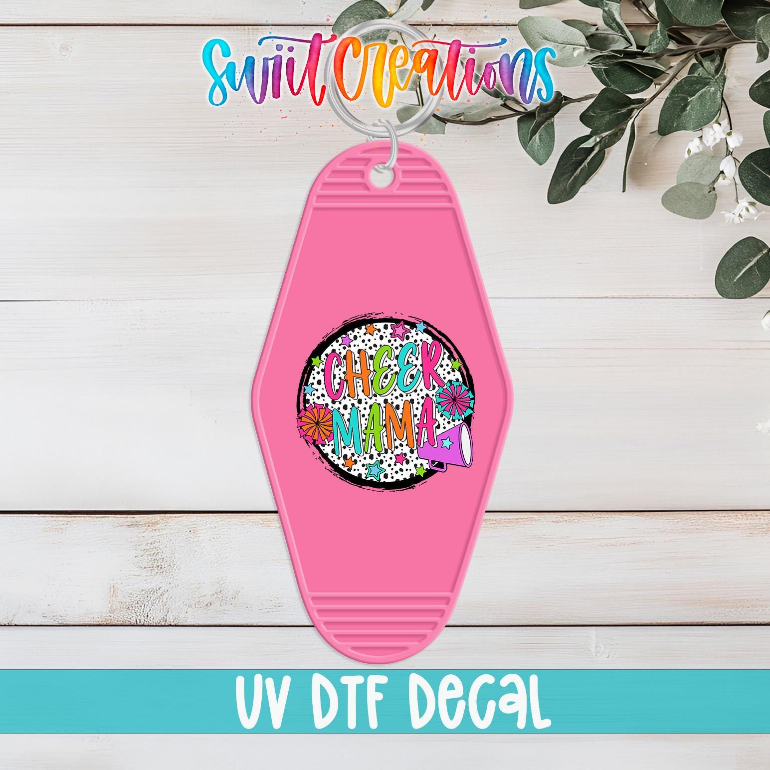 A pink keychain with a colorful, abstract design and the text "CHEER MAMA" printed on it, hanging on a wooden surface.