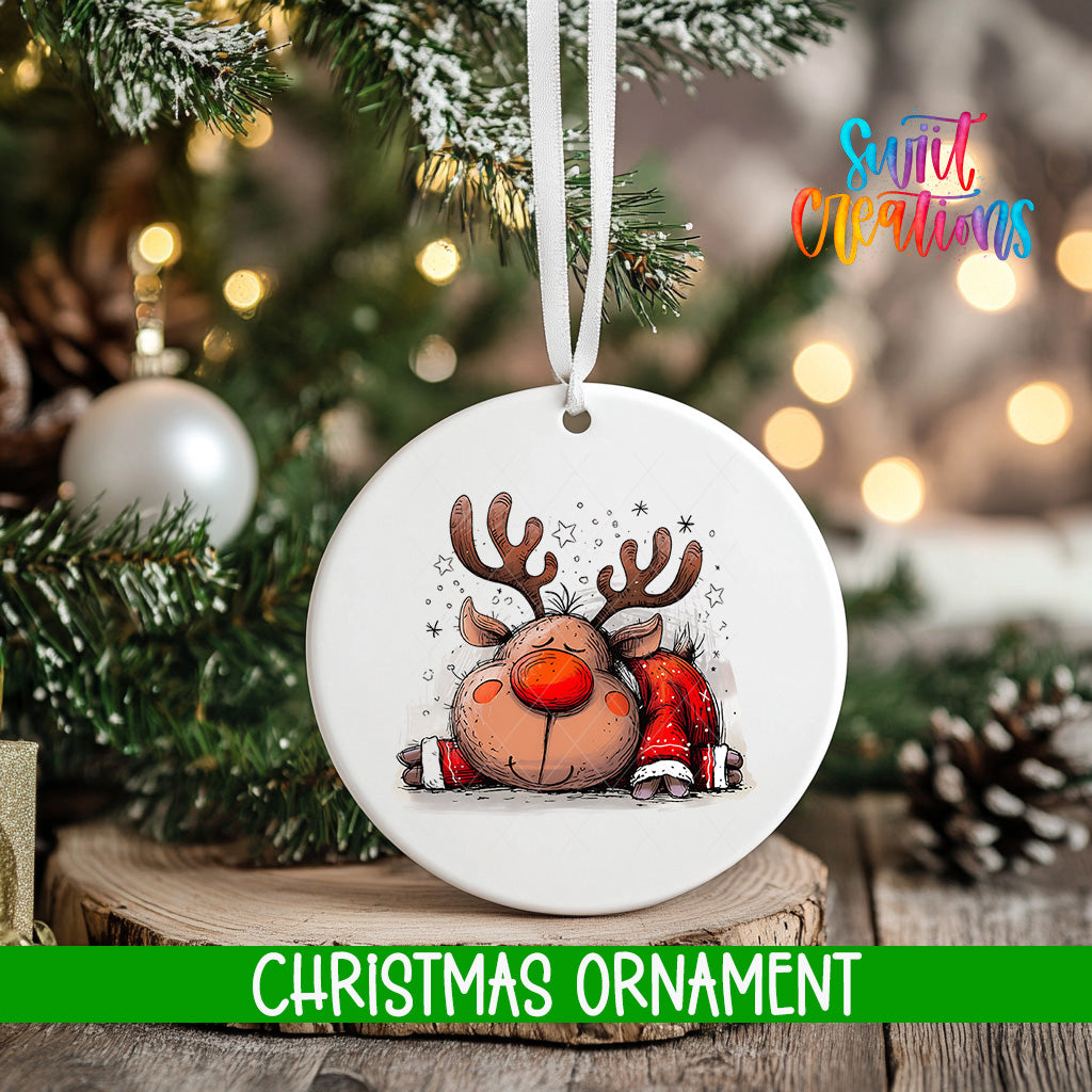 A white Christmas ornament with a cartoon reindeer design hangs on a tree branch, surrounded by pine cones and other holiday decorations.