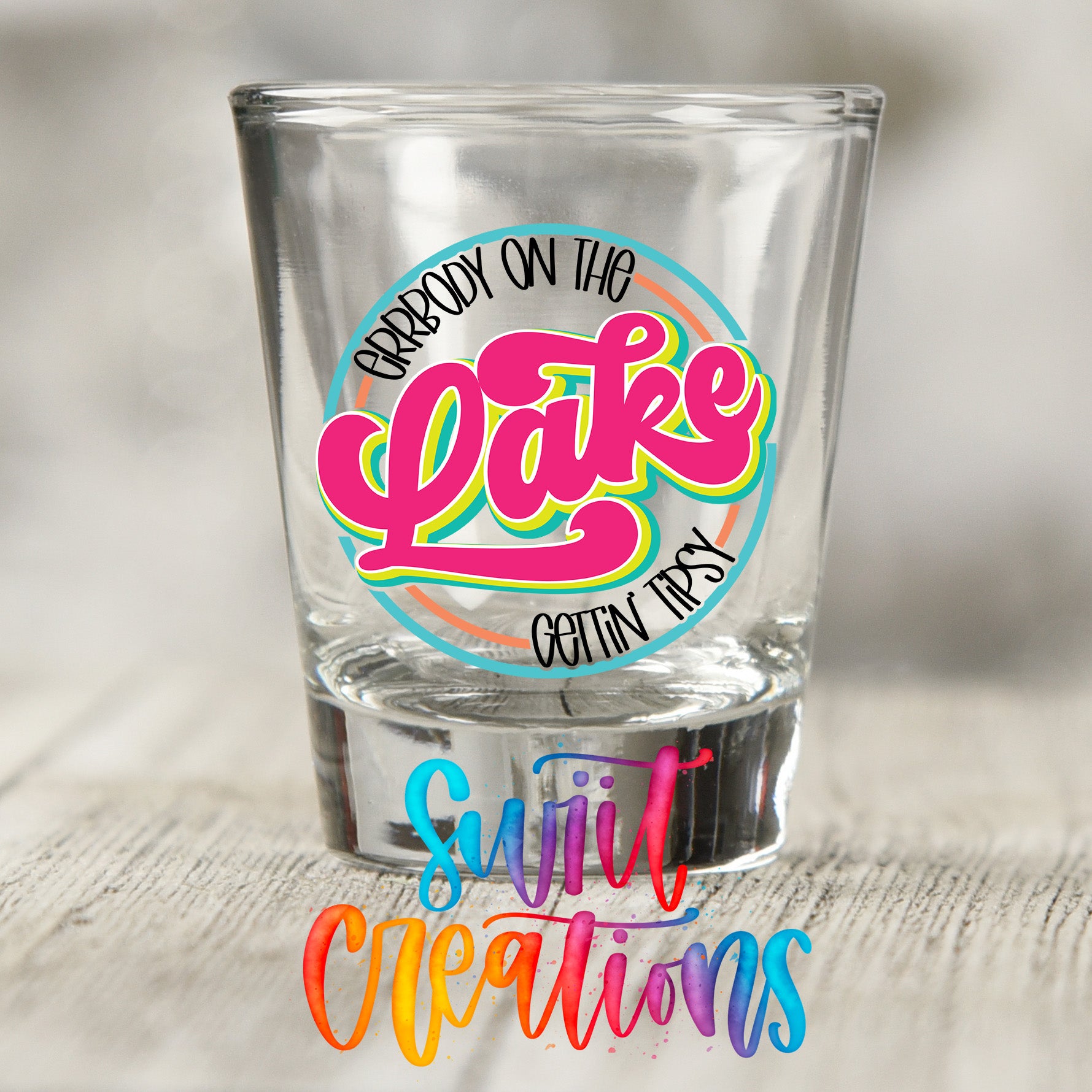 A clear shot glass with a colorful design and text on it, placed on a wooden surface.