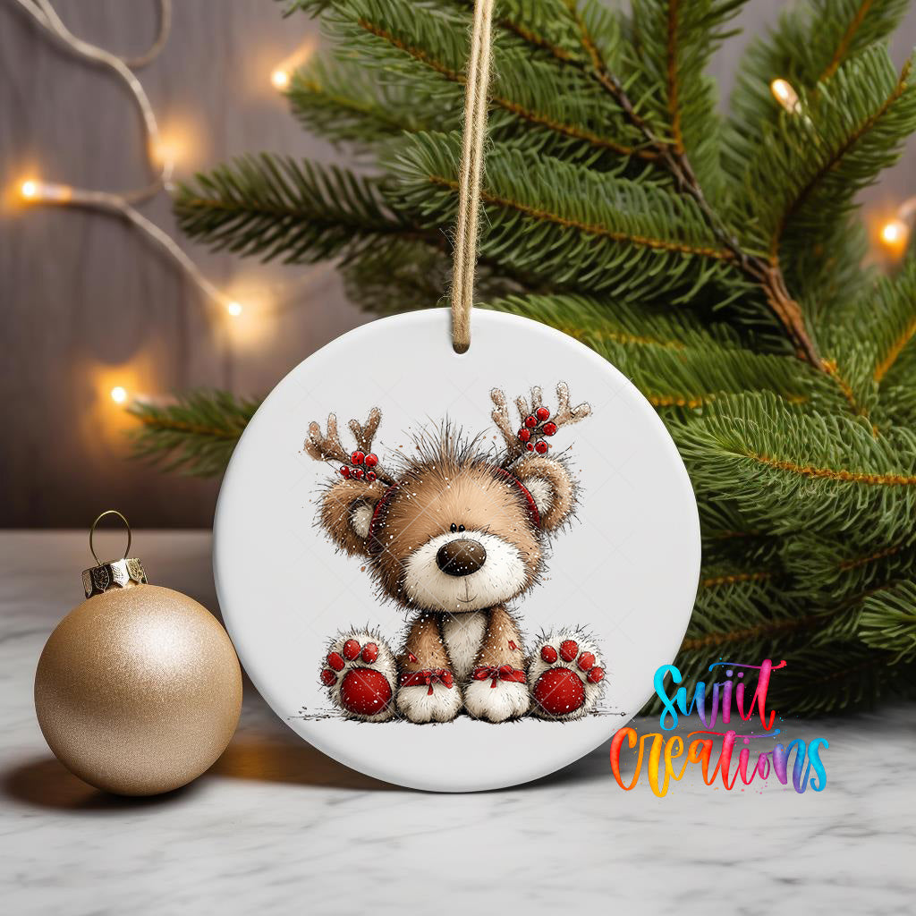 A white ceramic ornament featuring a teddy bear with antlers is displayed on a marble surface, with a Christmas tree and a gold ornament in the background.