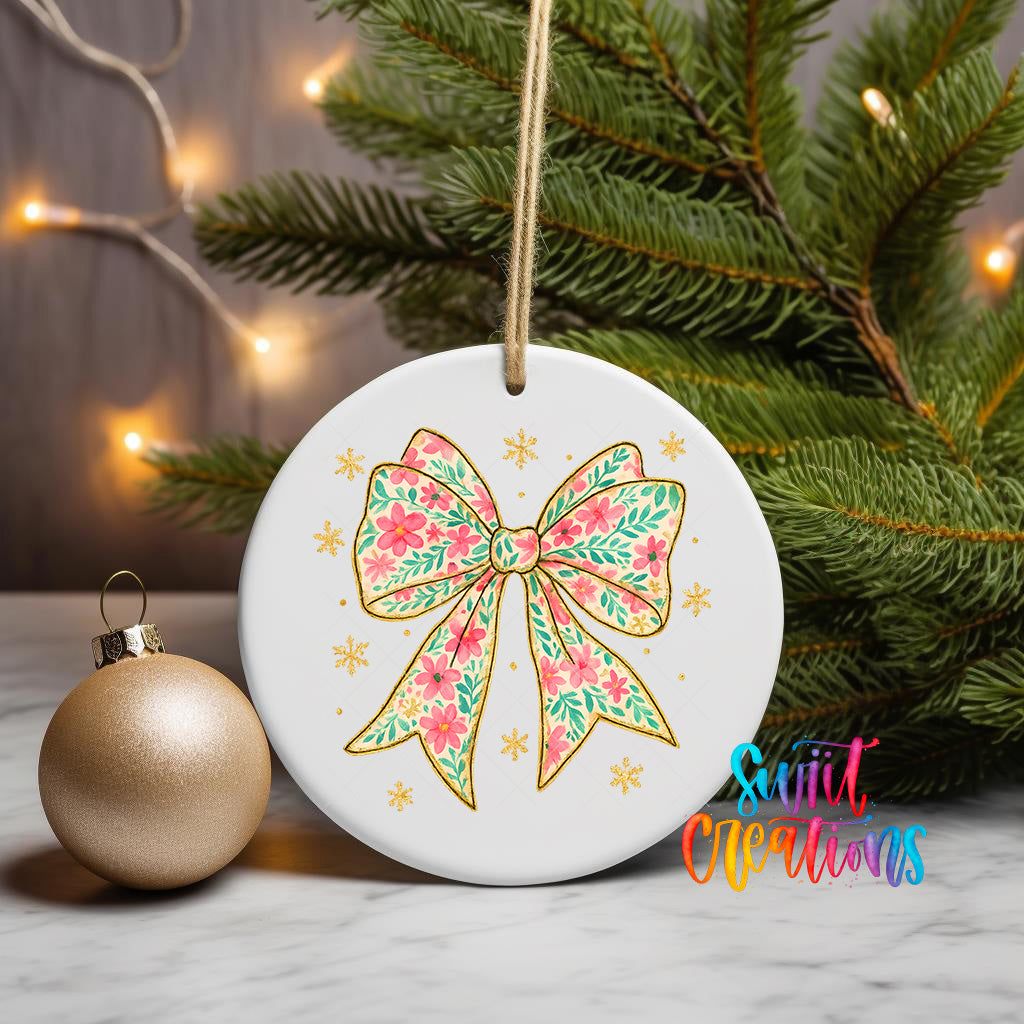 A white ceramic ornament with a floral bow design and snowflake decorations is displayed on a marble surface, accompanied by a gold ornament and a pine tree with twinkling lights in the background.