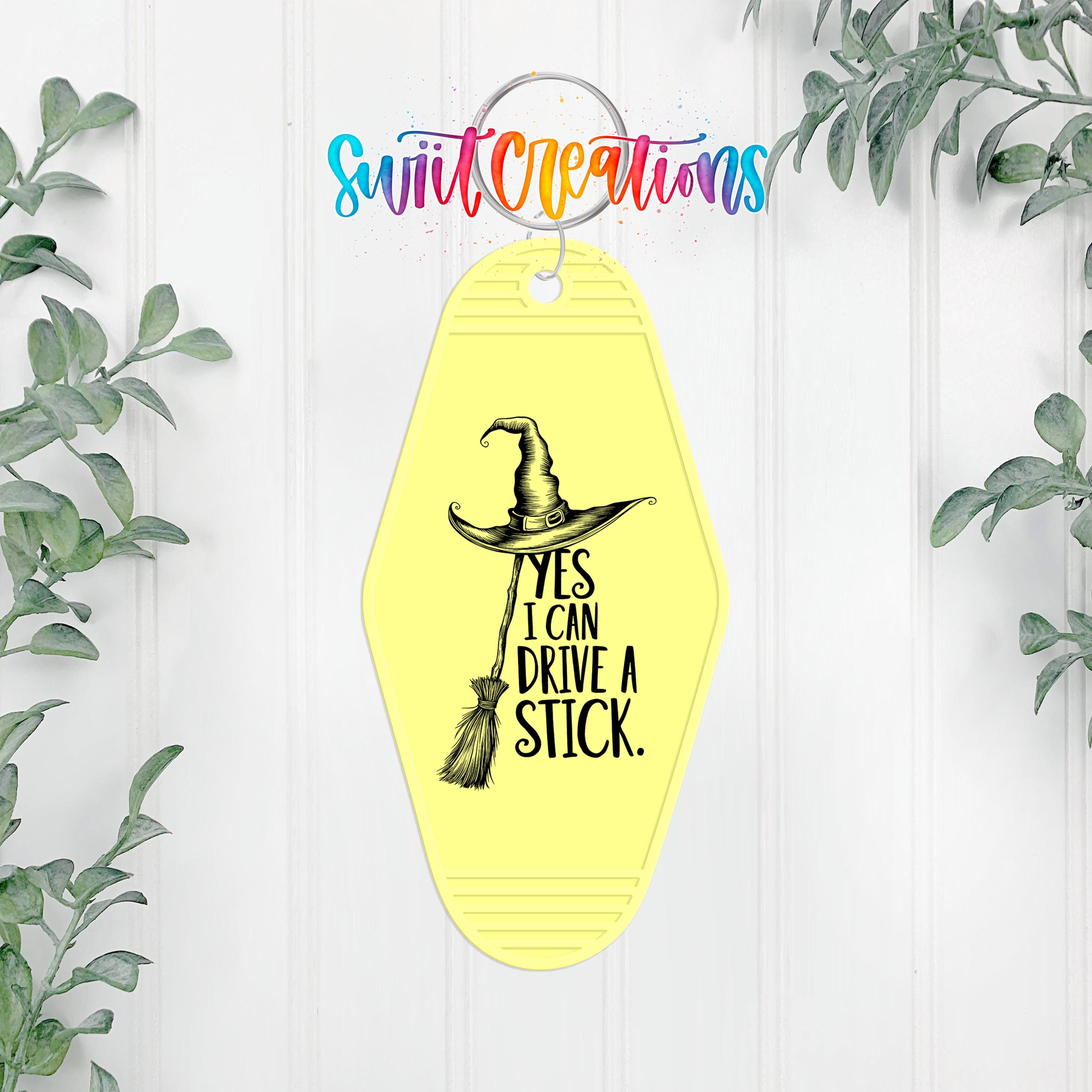 A yellow keychain with a witch's broom and the text "YES I CAN DRIVE A STICK" printed on it.