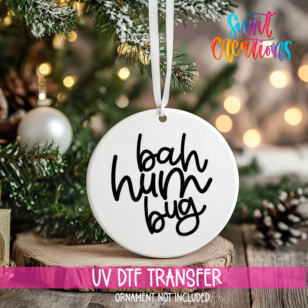 A white ornament with the text "bah hum bug" hanging on a Christmas tree, with a blurred background of pine branches and twinkling lights.