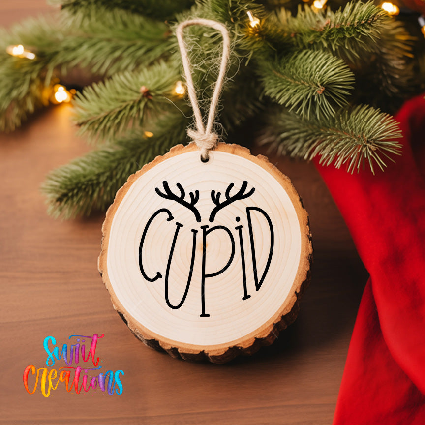 A wooden ornament with the word "CUPID" written on it, hanging on a Christmas tree with pine branches and twinkling lights.
