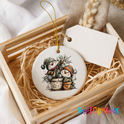 A ceramic ornament featuring two snowmen in a pot, sitting on a bed of straw. The ornament is placed in a wooden crate, and there is a tag attached to it.