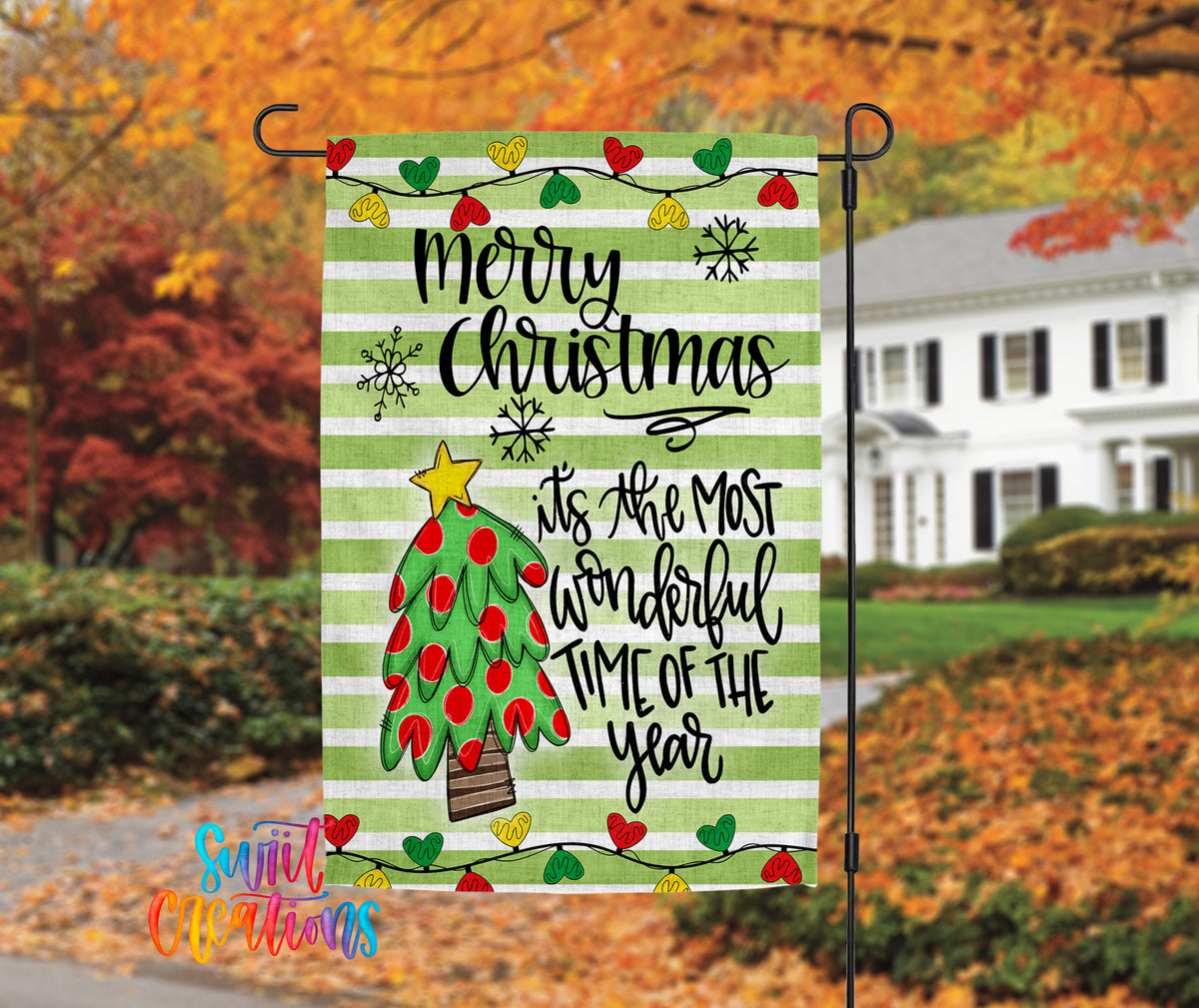 A festive Christmas-themed flag is displayed in front of a house with autumn foliage in the background.
