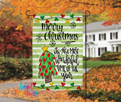 A festive Christmas-themed flag is displayed in front of a house with autumn foliage in the background.