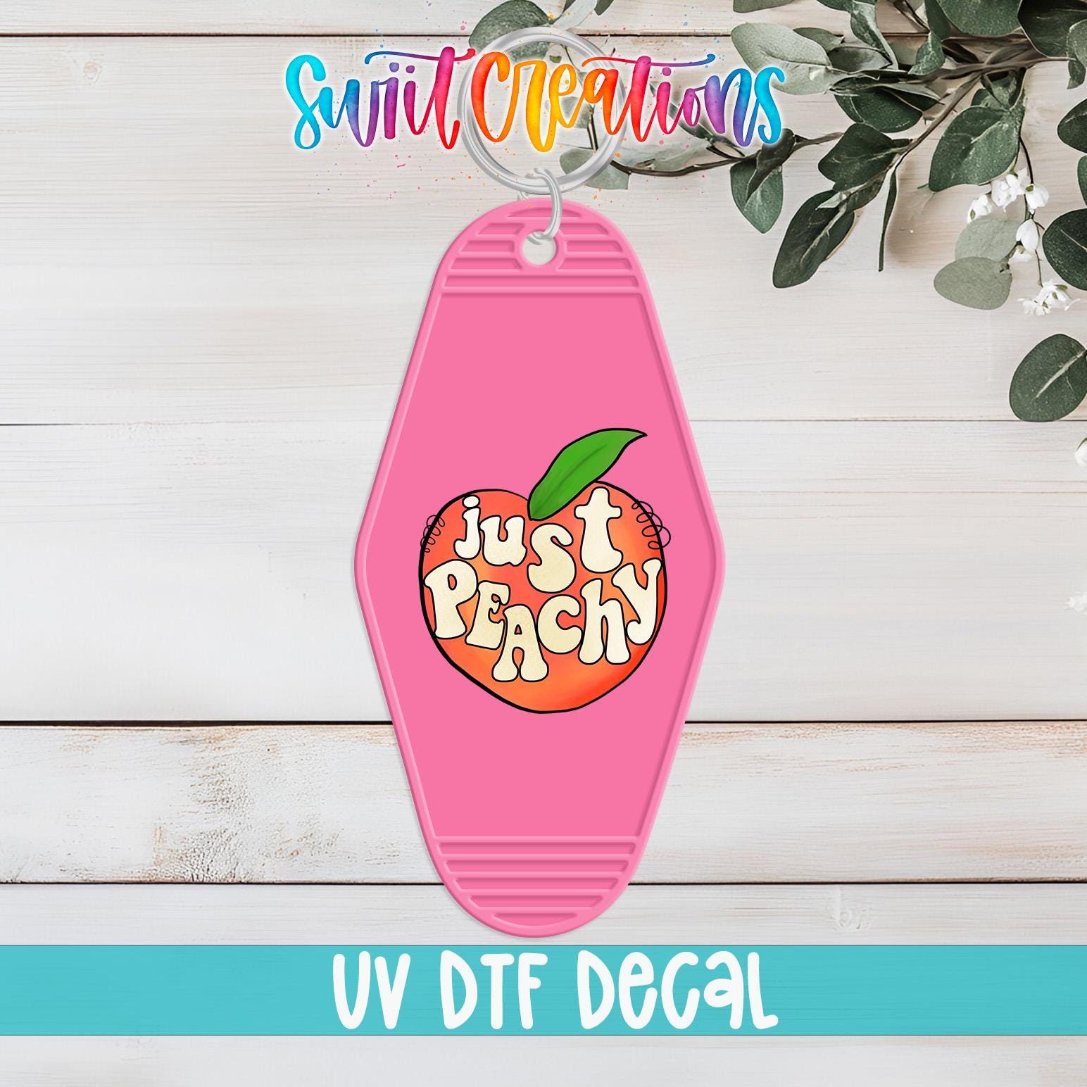 A pink keychain with a peach design and the text "Just Peachy" printed on it, placed on a wooden surface with some greenery in the background.