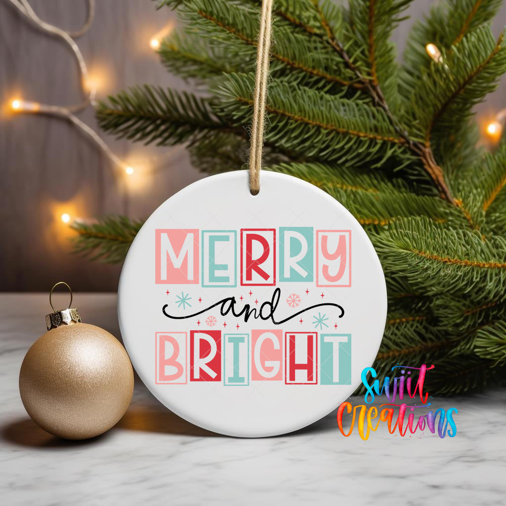 A white ceramic ornament with the text "MERRY AND BRIGHT" in pink, blue, and red colors, hanging from a string. The ornament is placed next to a gold ornament and a Christmas tree, creating a festive holiday scene.