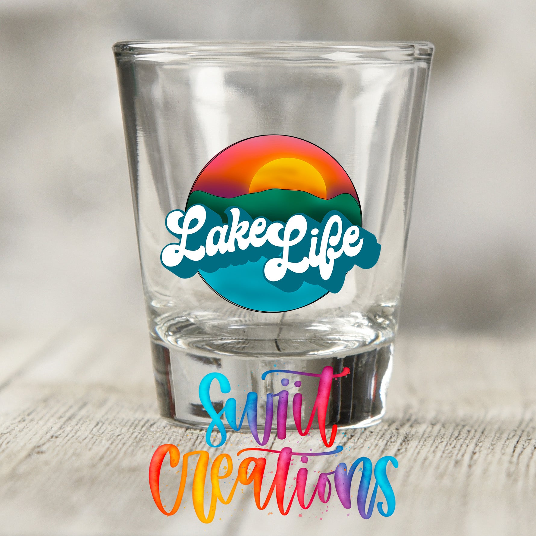 A clear shot glass with a colorful graphic design on it, featuring the text "LakeLife" and "Suit Creations" written in a stylized font.