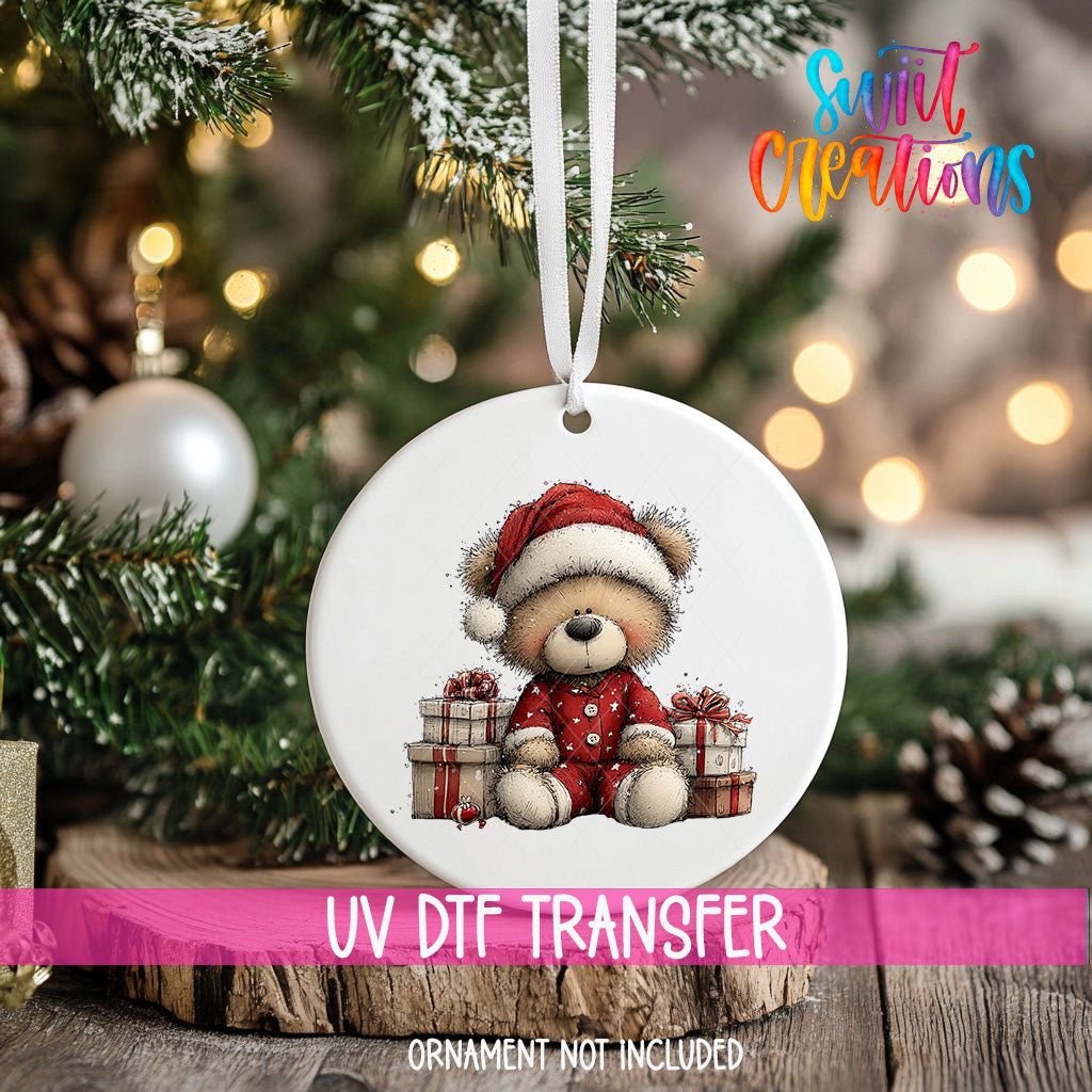 A white ornament with a teddy bear wearing a Santa hat and holding presents is hanging on a Christmas tree.