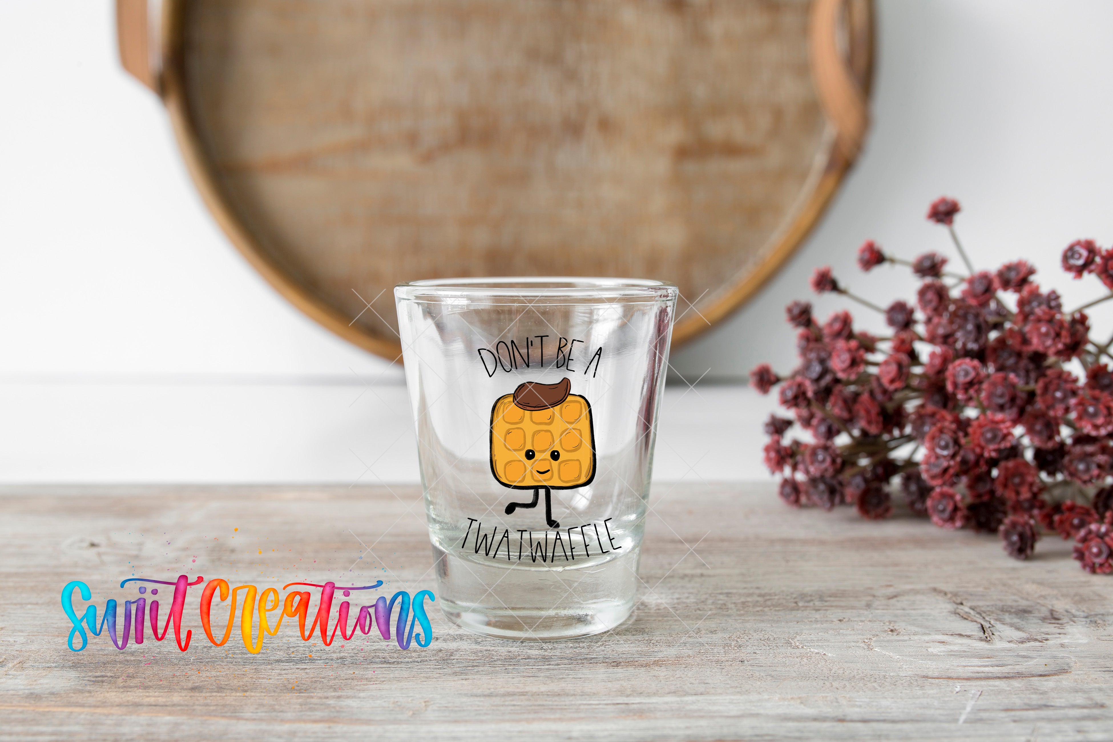 A shot glass with a cartoon character on it, placed on a wooden surface next to a bunch of red flowers.