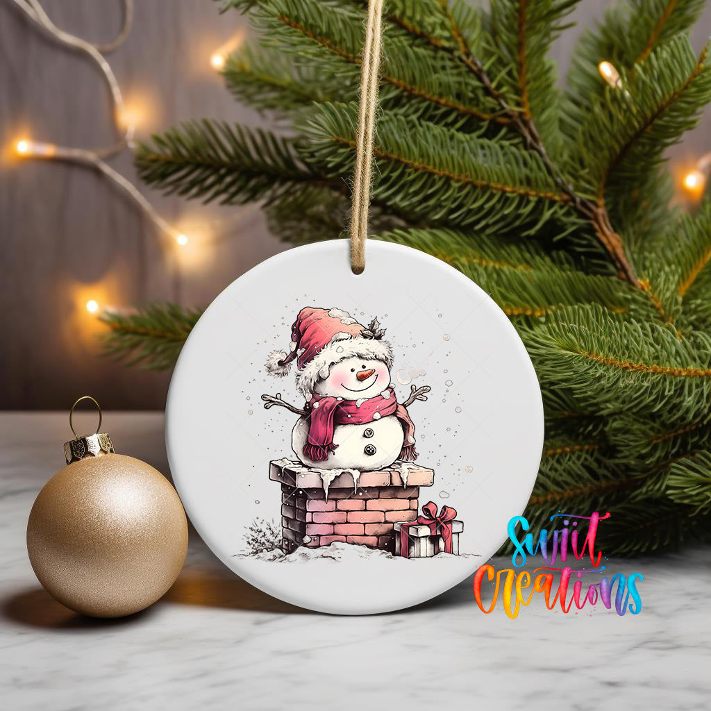 A white ceramic ornament featuring a snowman sitting on a brick chimney, surrounded by Christmas decorations and a gold ornament on a marble surface.