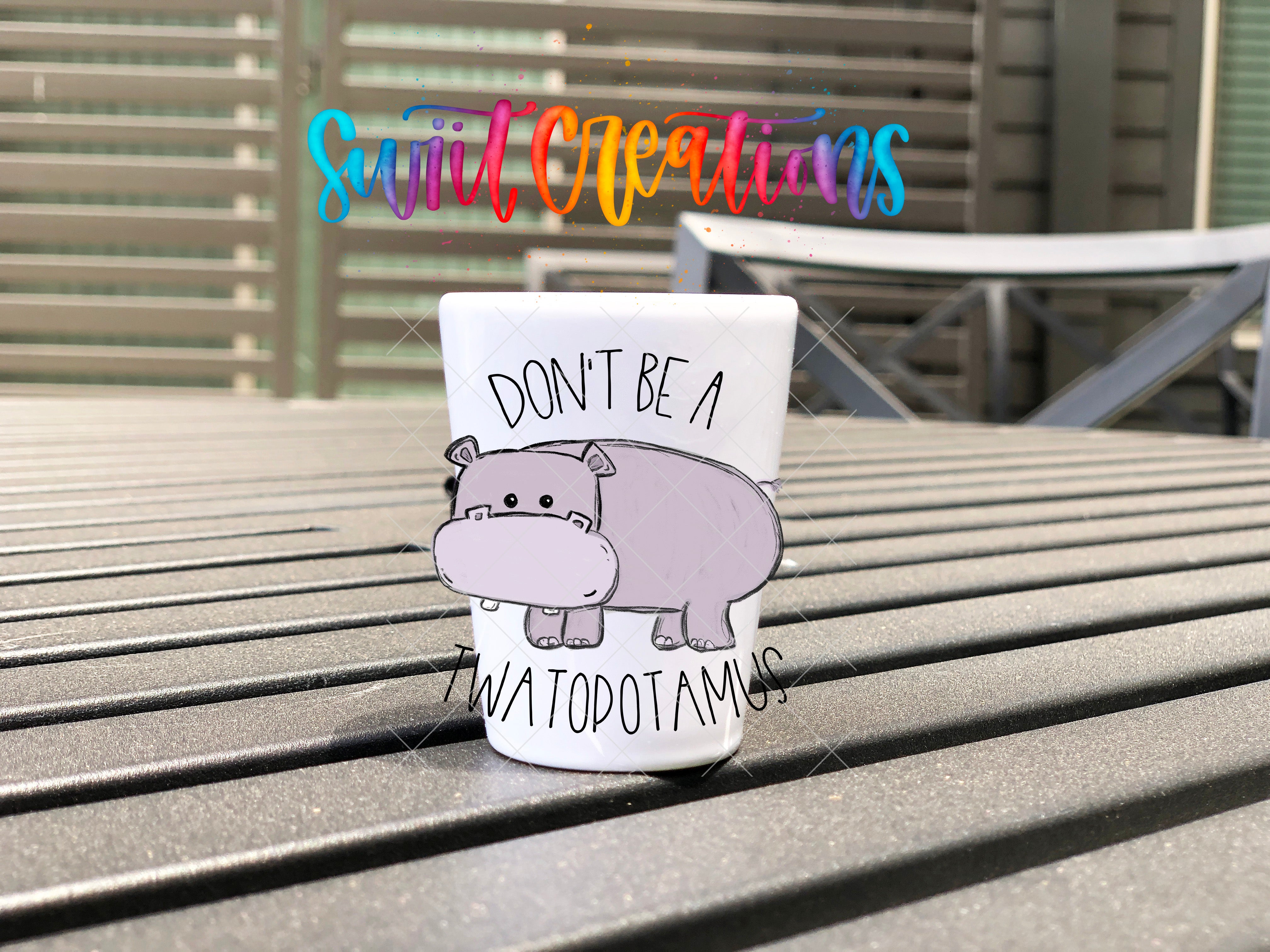 A white mug with a cartoon image of a hippopotamus and the text "DON'T BE A TWATOPOTAMUS" printed on it, placed on a wooden surface.