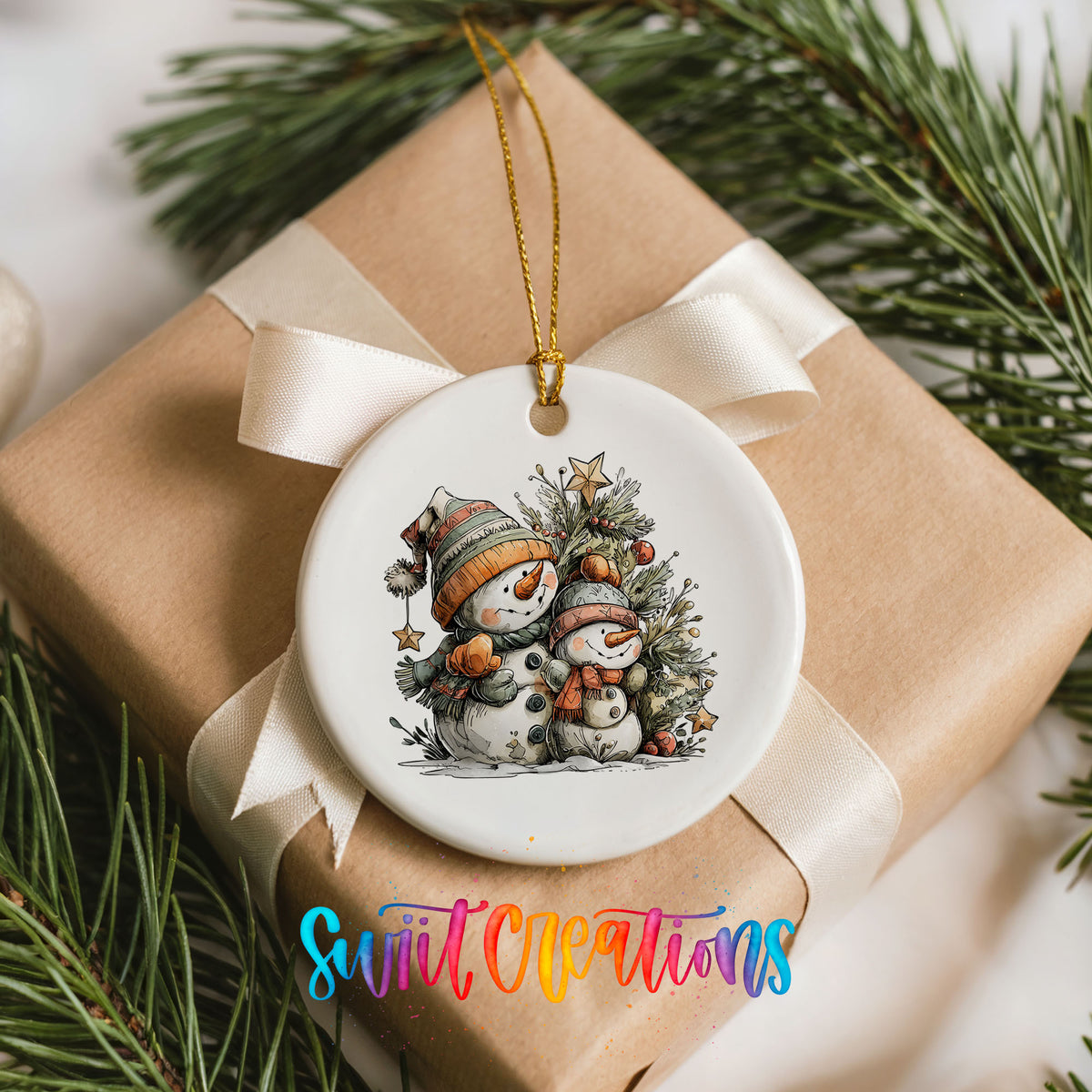A white ceramic ornament with a snowman and pine tree design hangs from a ribbon on a brown gift box, surrounded by pine branches.