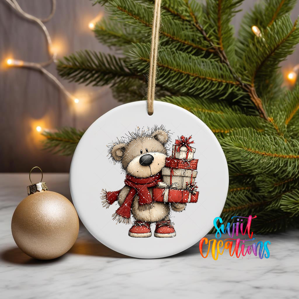 A white ceramic ornament featuring a cartoon teddy bear holding a gift bag, placed on a marble surface next to a gold ornament and a Christmas tree.