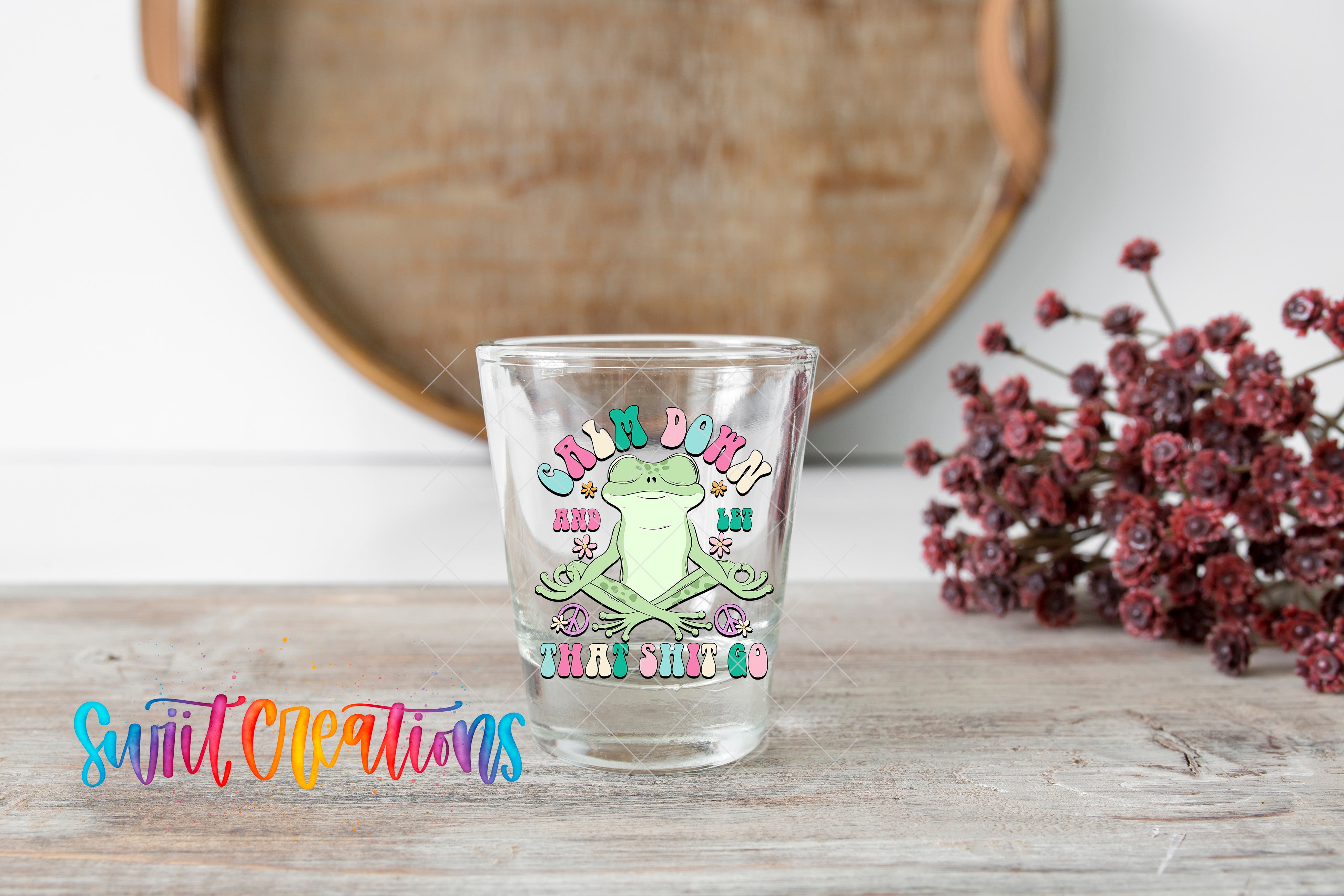 A glass with a colorful frog design and text on it, placed on a wooden surface next to a bunch of red flowers.