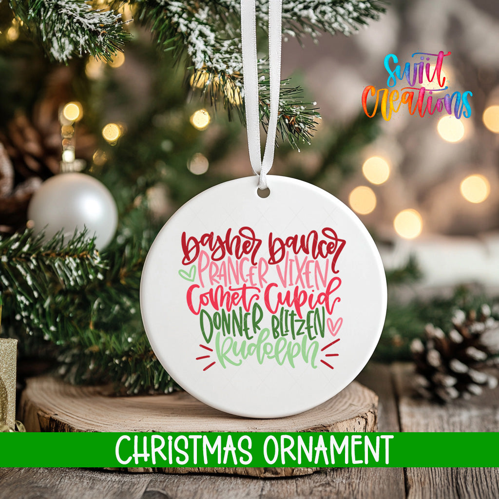 A white Christmas ornament with a colorful design hangs on a tree branch, surrounded by pine cones and other holiday decorations.