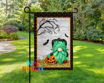 A Halloween-themed garden flag featuring a green gnome holding a jack-o-lantern, with a spooky tree and bats in the background.
