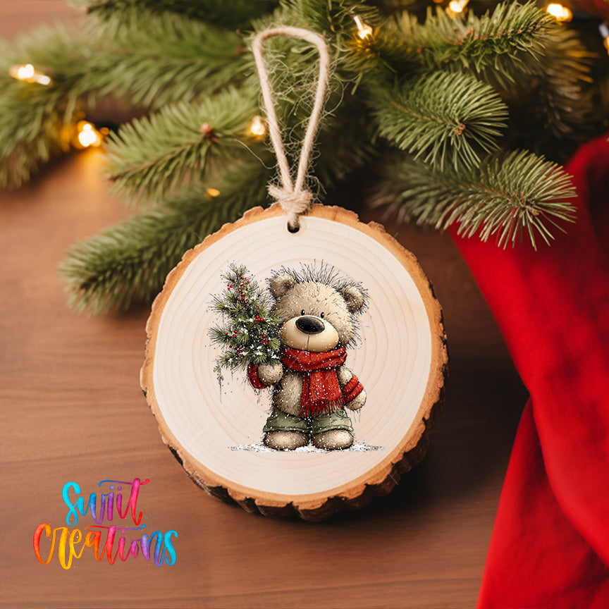 A wooden ornament featuring a teddy bear holding a Christmas tree, hanging on a Christmas tree with lights visible in the background.