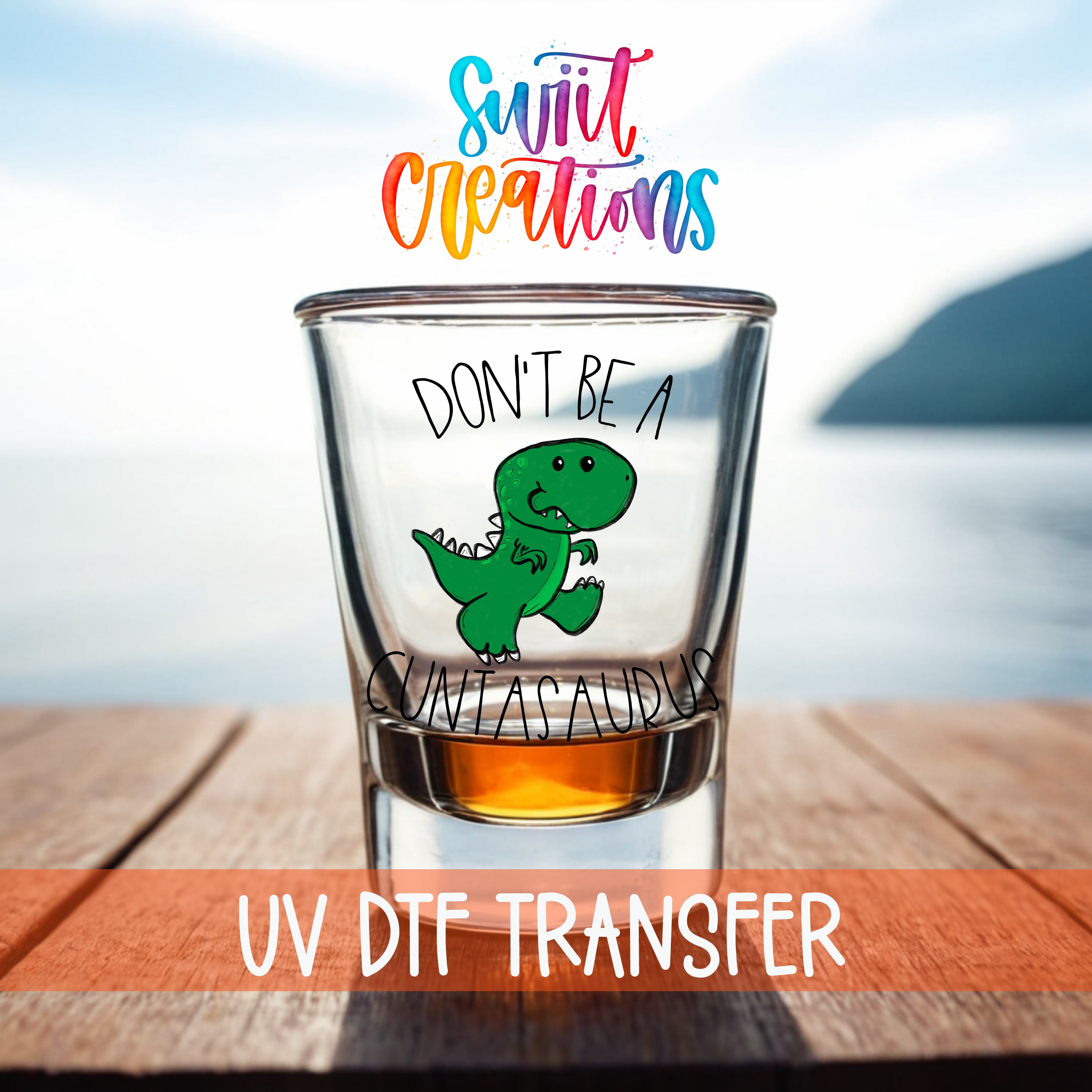 a shot glass with a cartoon dinosaur and text on it, placed on a wooden surface with a body of water in the background.