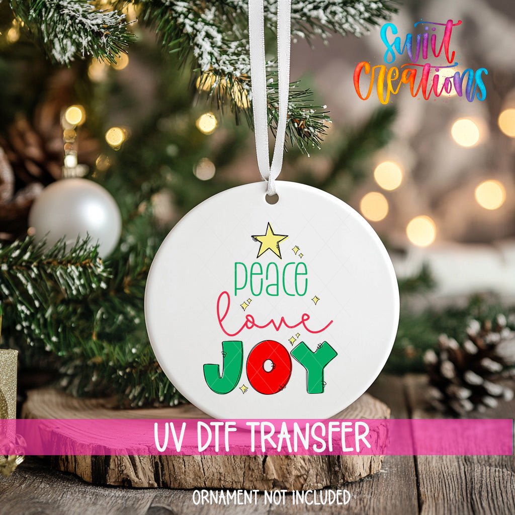 A white ornament with a message "Peace love joy" is hanging on a Christmas tree, with a blurred background of lights and greenery.