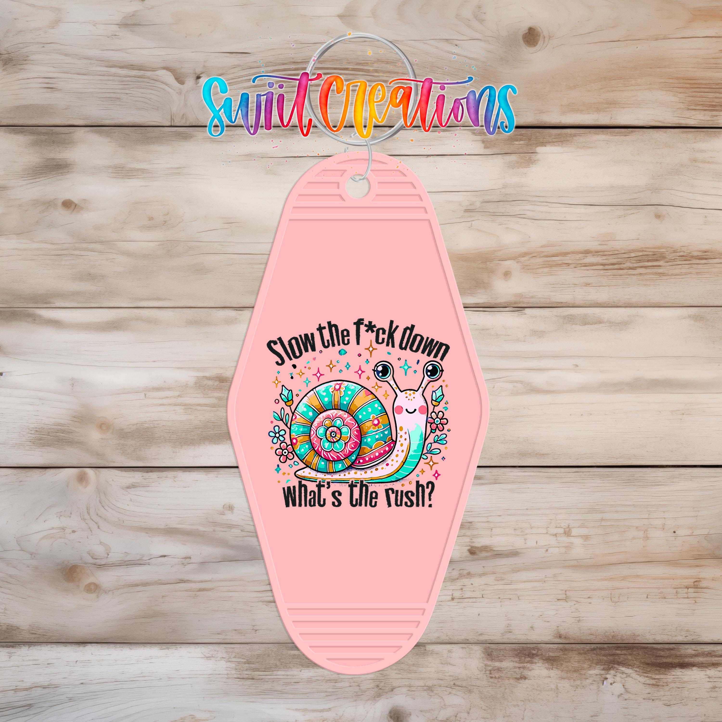 A pink keychain with a cartoon snail and the text "Slow the fuck down what's the rush?"