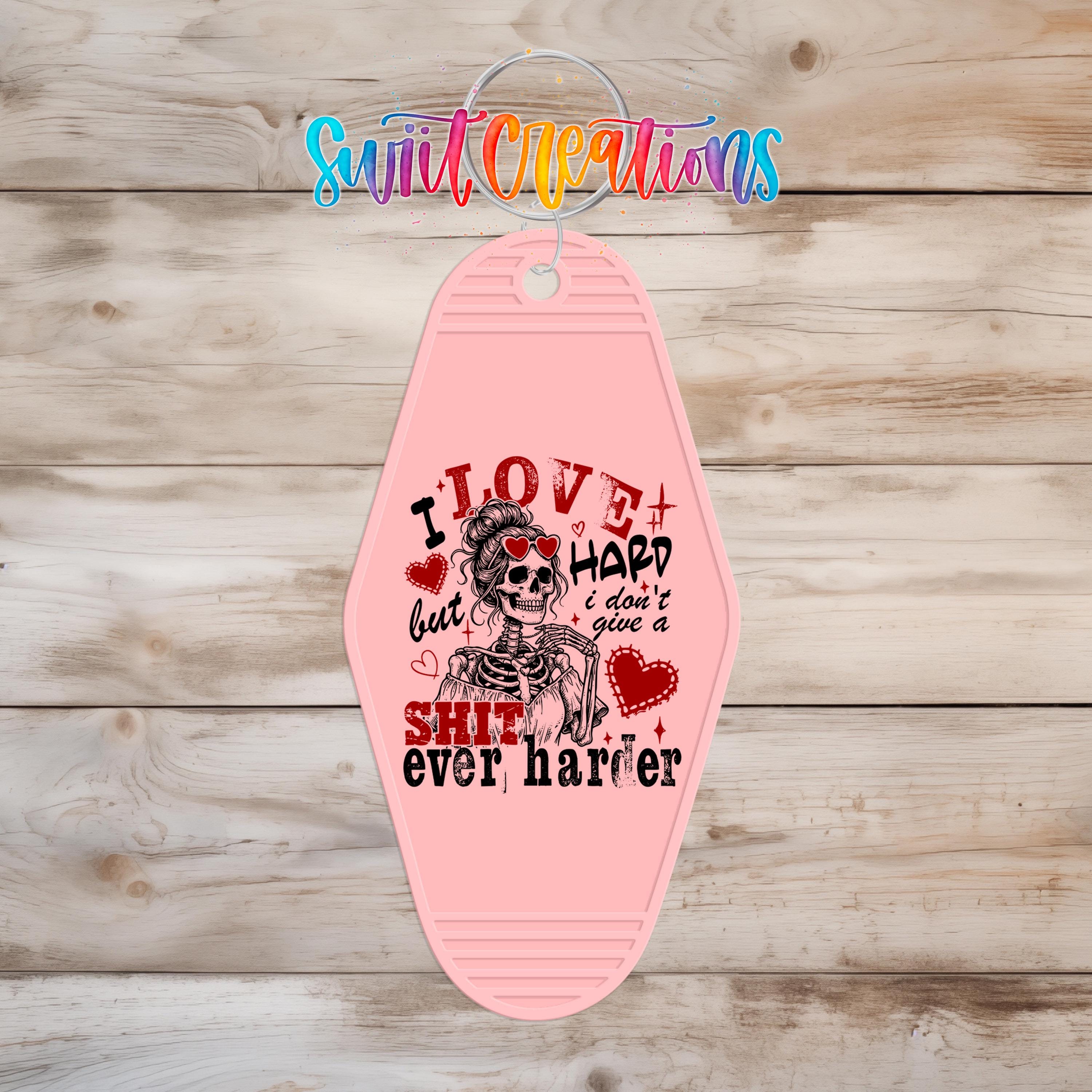 A pink keychain with a skull design and text that says "I LOVE HARD but I DON'T GIVE A SHIT ever harder".