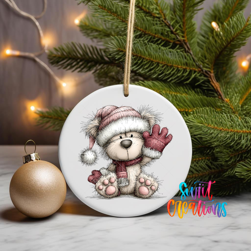 A white ceramic ornament featuring a teddy bear wearing a red hat and scarf, with a Christmas tree in the background and a gold ornament on a marble surface.