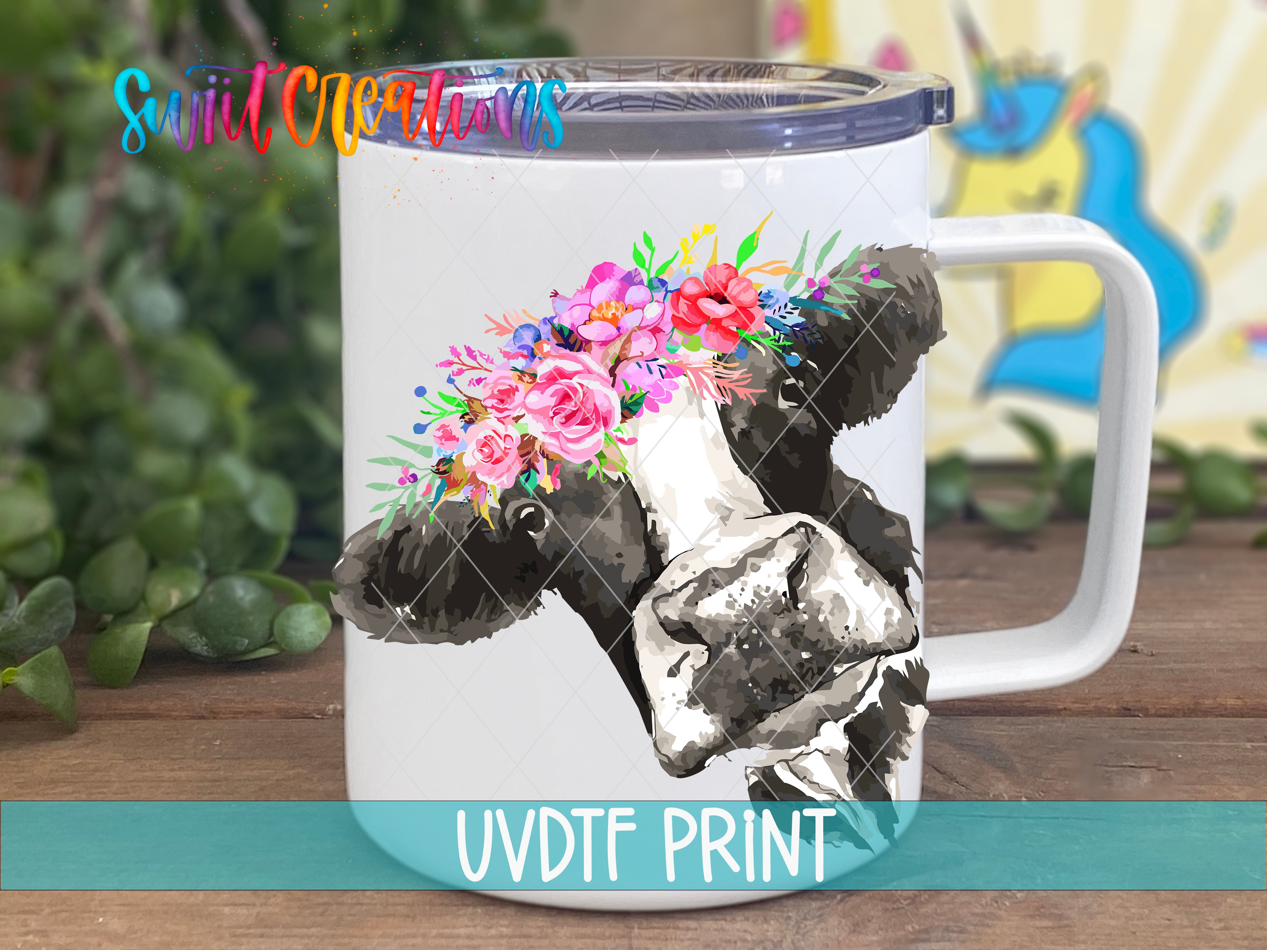 A white mug with a black and white cow design and a floral wreath on its head, placed on a wooden surface.