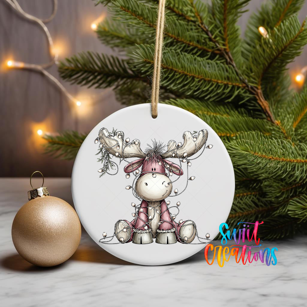 A white ornament with a cartoon moose and Christmas lights is displayed on a table, with a gold ornament and a pine tree in the background.