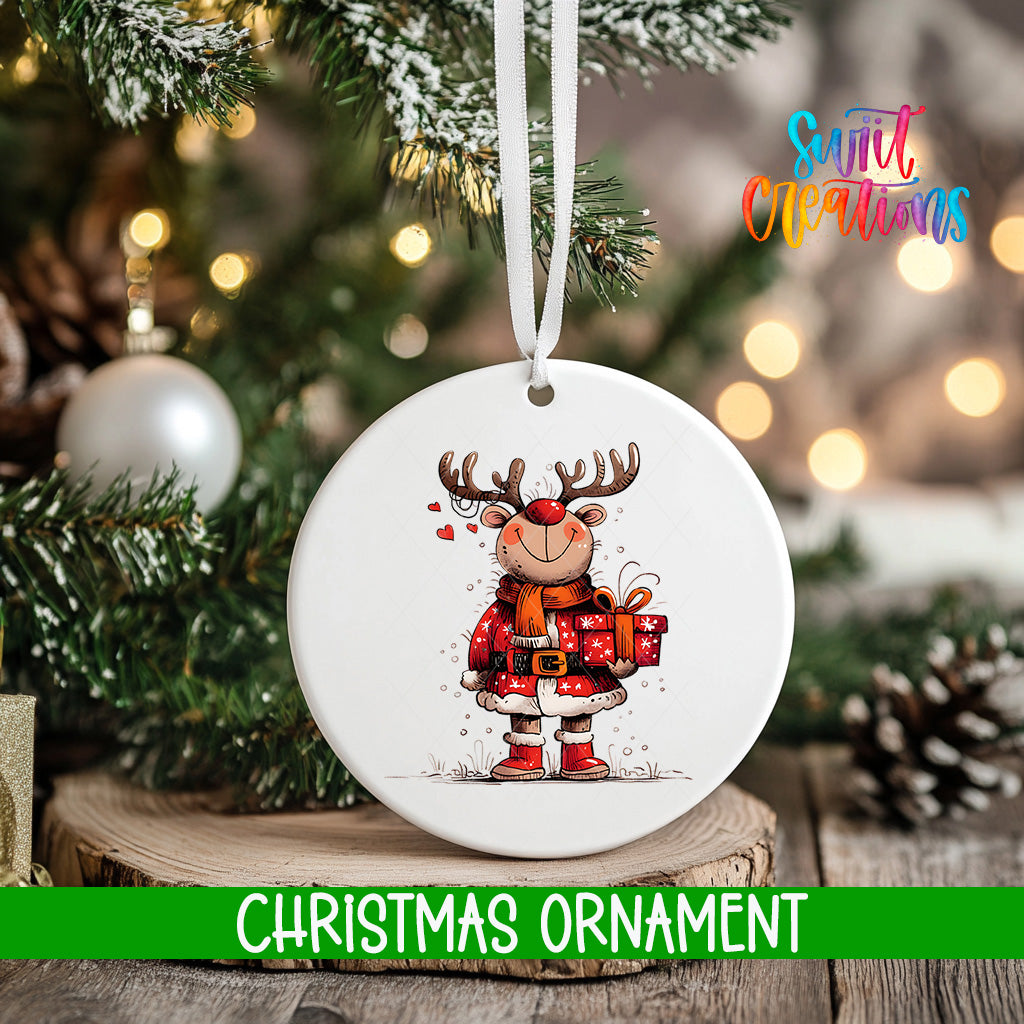 A white Christmas ornament featuring a cartoon reindeer holding a gift, hanging on a tree branch with a blurred background of lights and greenery.