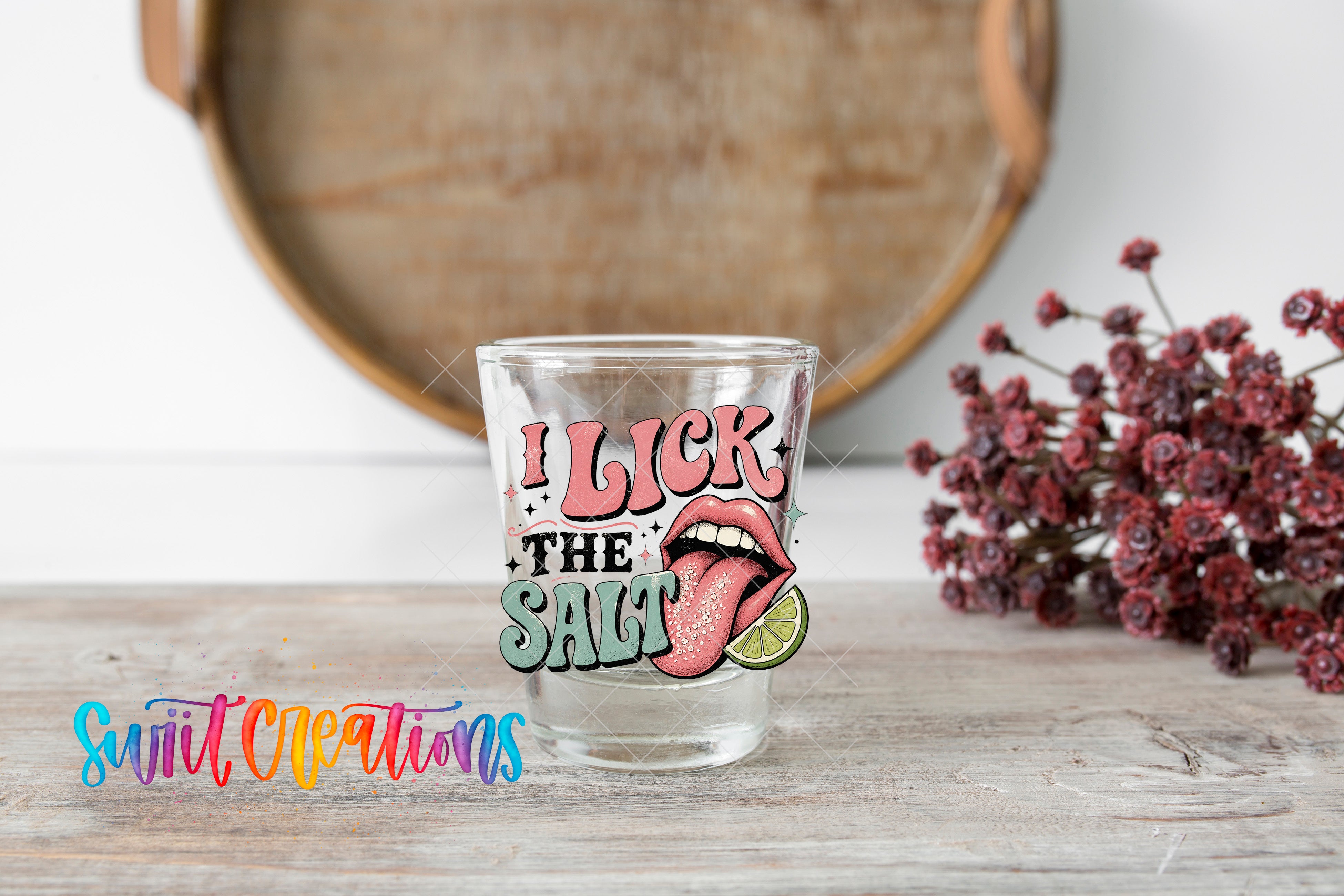 A glass with a colorful graphic design featuring a tongue sticking out and the text "I LICK THE SALT" on it, placed on a wooden surface.