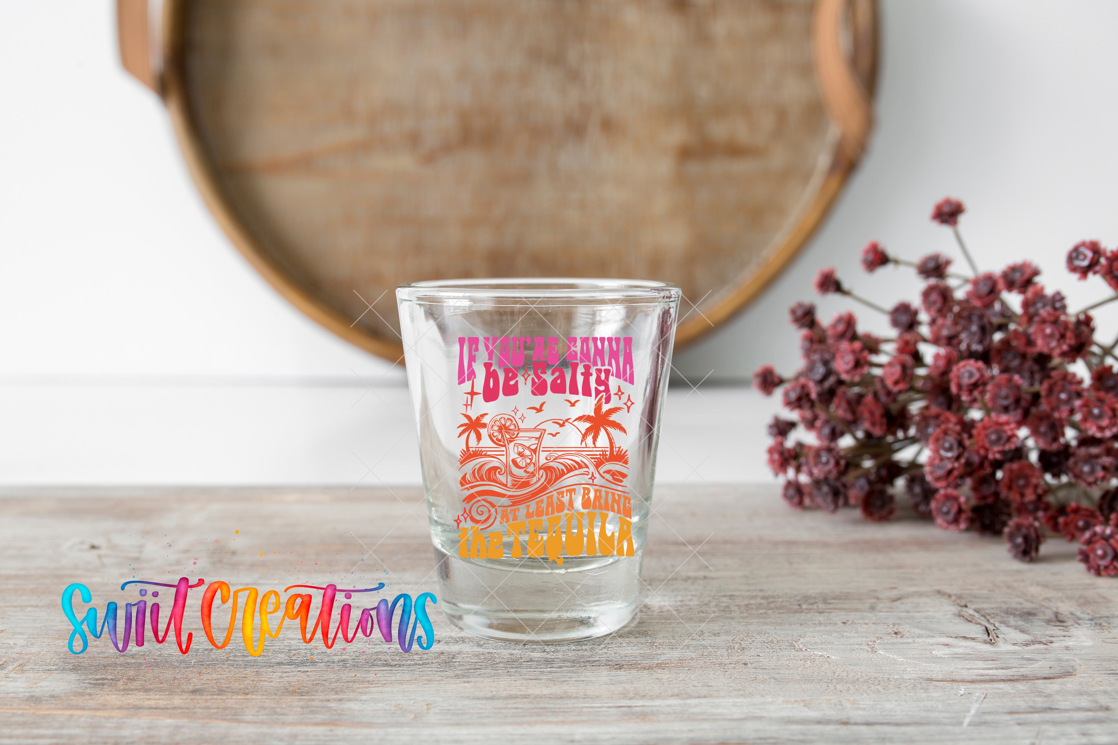 A shot glass with a colorful design sits on a wooden surface, with a bouquet of flowers and a wooden tray in the background.