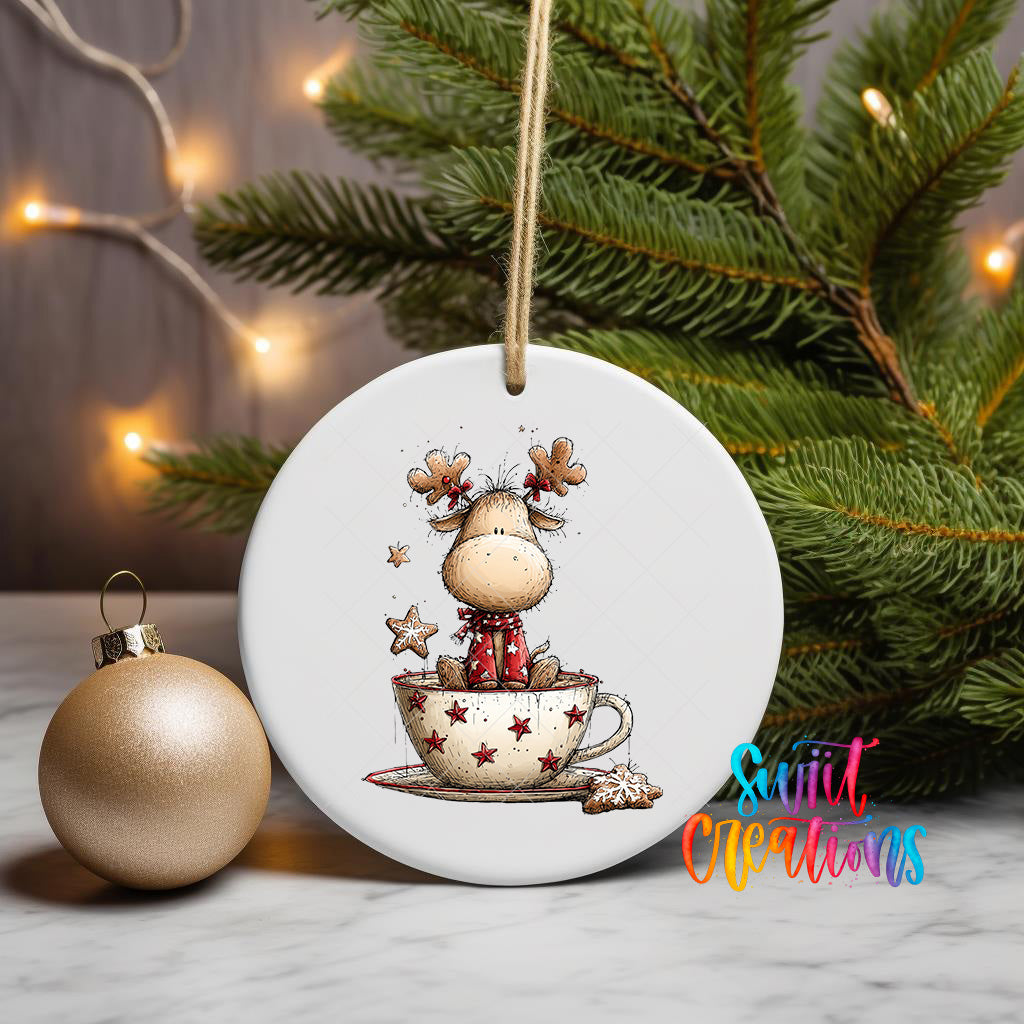 A white ceramic ornament featuring a cartoon reindeer sitting in a cup of coffee, surrounded by stars, is displayed on a table with a Christmas tree and a gold ornament in the background.
